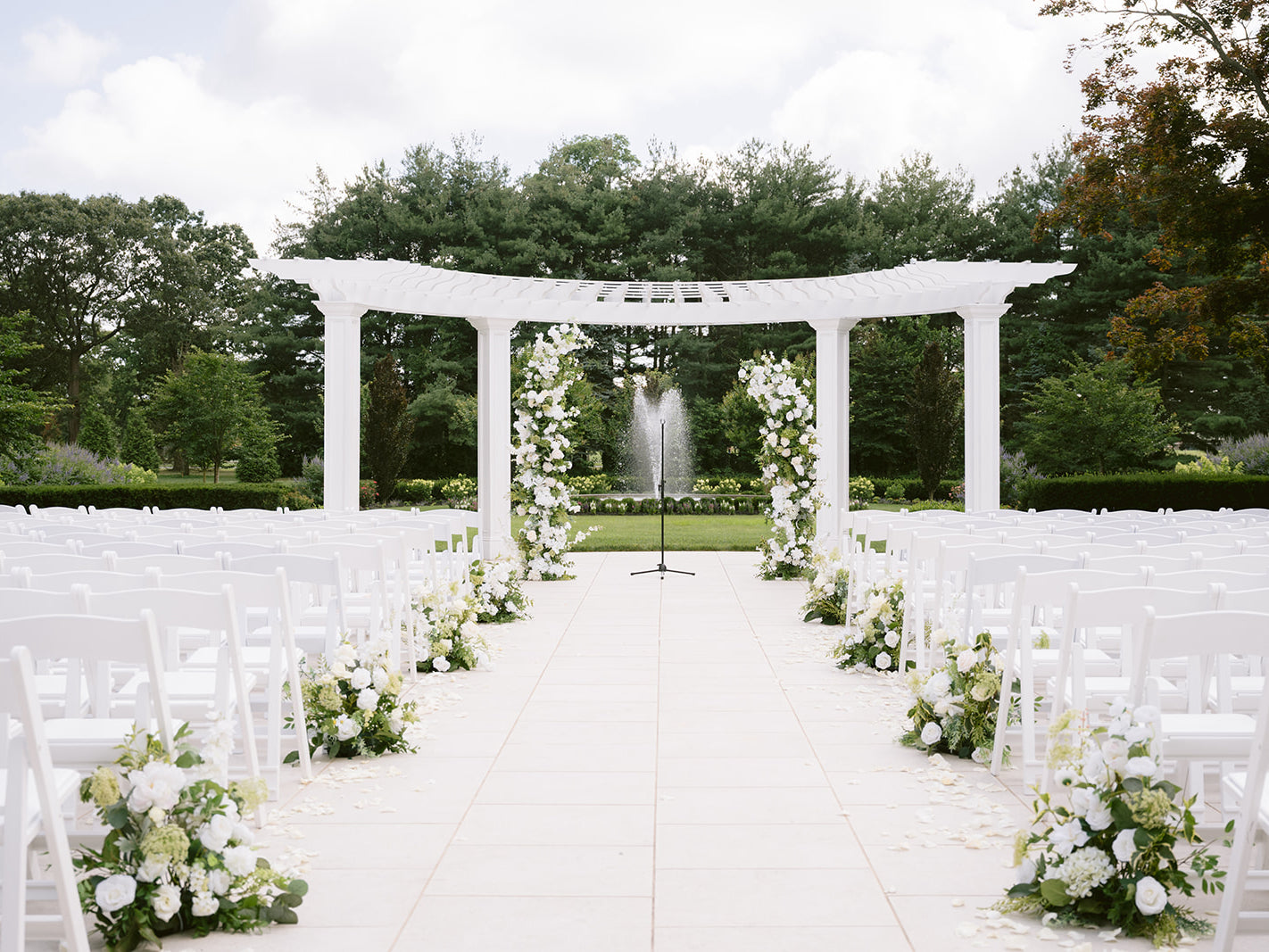 Outdoor wedding ceremony setup with white chairs, floral arrangements, and a white archway.