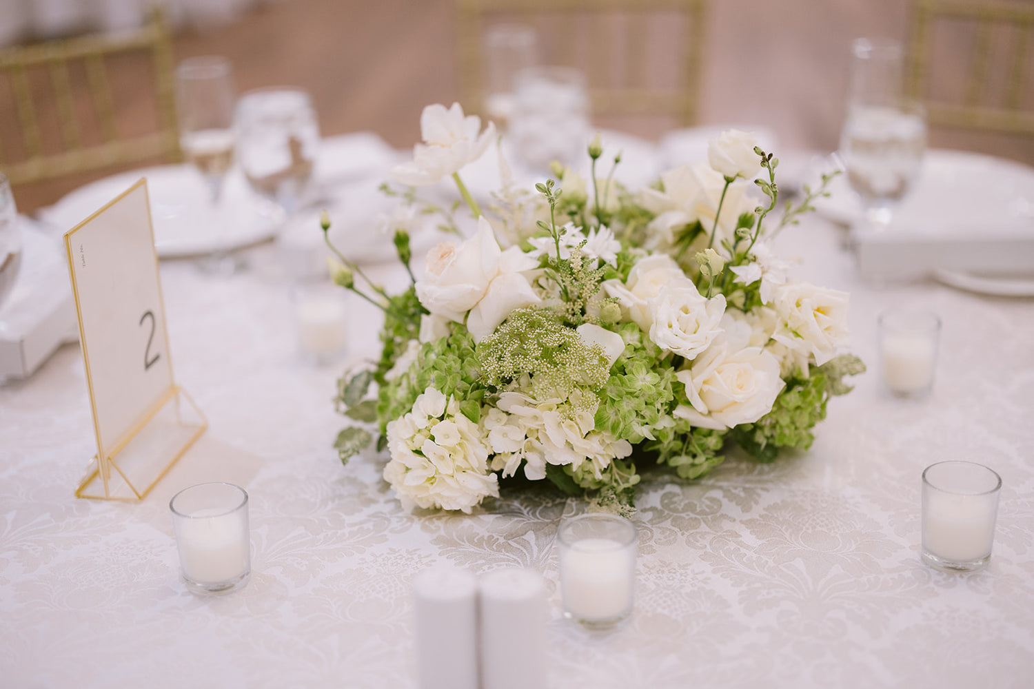 Floral centerpiece with white and green flowers on a table with candles and a table number.