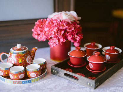 Tea set with red teacups and a colorful teapot on a wooden tray, accompanied by pink flowers.