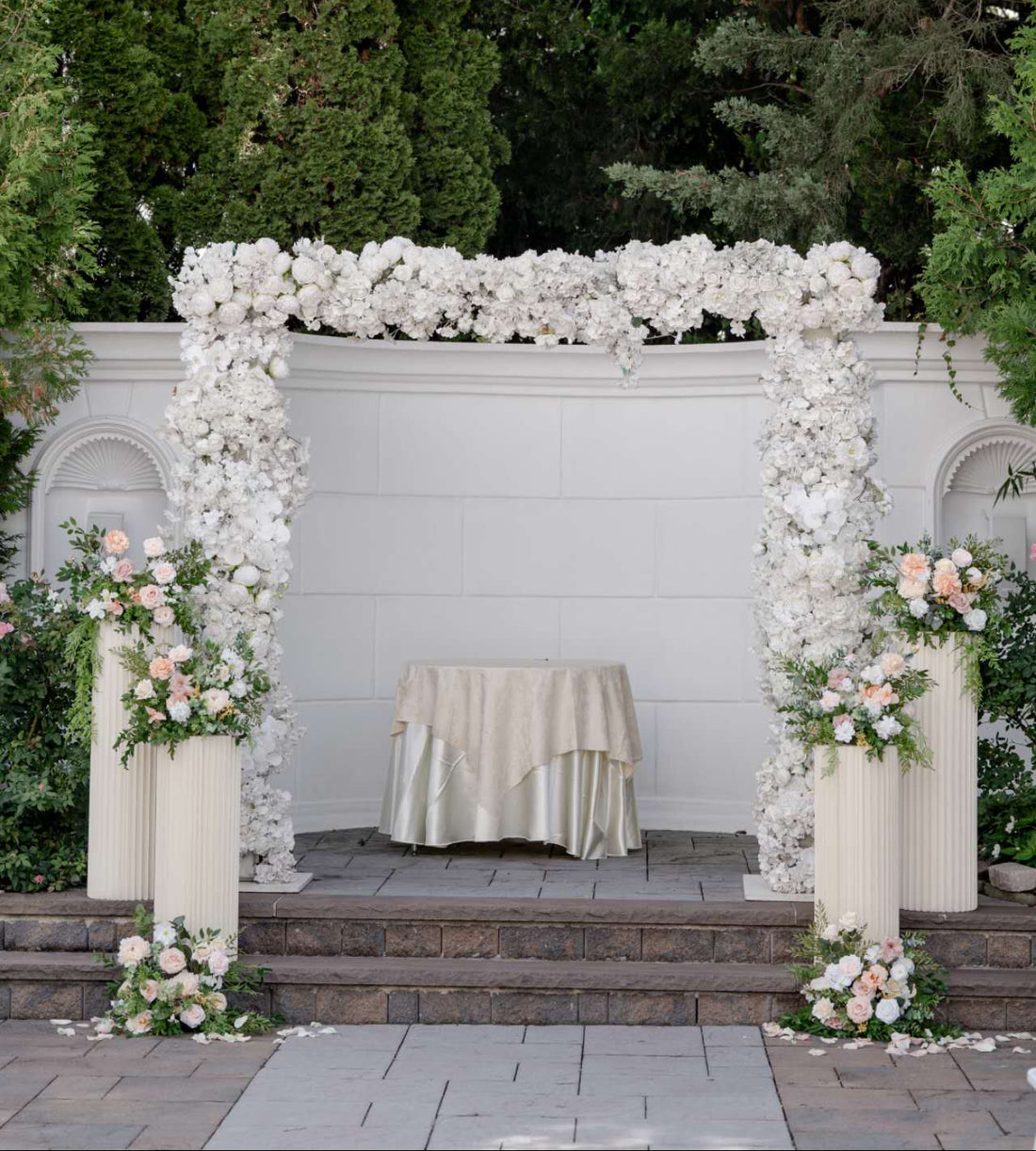 Decorative arch with white flowers and greenery in front of a white wall.