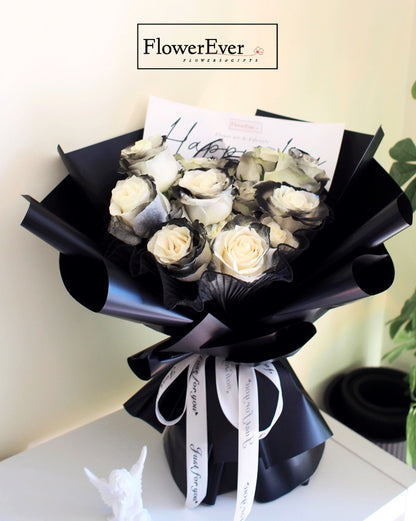 Sixteen brilliant white preserved rose blooms and black knight rose buds artfully arranged in a round bouquet wrapped with sleek black gift paper.