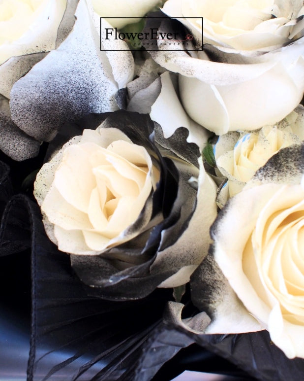 A round floral arrangement featuring 16 vivid white preserved roses and deep black rose buds bundled in black wrapping paper.