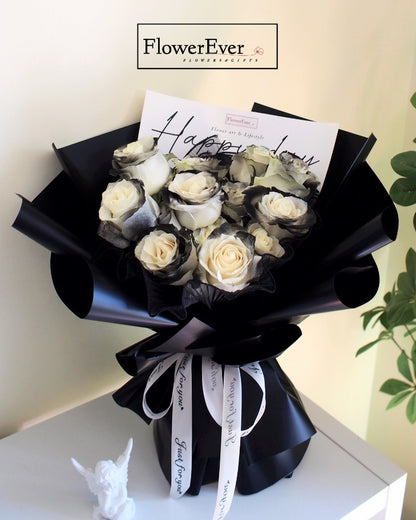 Sixteen stems including preserved white roses and black knight rose buds arranged in a Russian inspired round bouquet encased in black paper.