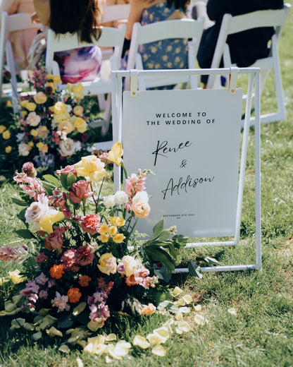 Wedding welcome sign with floral arrangement on a grassy outdoor setting