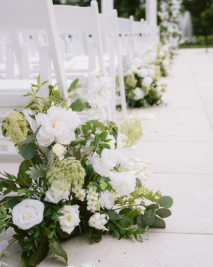 White flowers and greenery arranged along a white aisle with chairs.