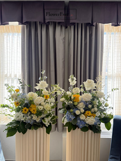 Two flower arrangements on stands in front of a window with curtains.